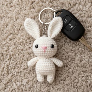 Peut inclure: Un porte-clés en forme de lapin blanc crocheté, avec des yeux noirs et un nez rose. Le lapin est attaché à un porte-clés argenté, à côté d'une clé de voiture noire. Le porte-clés est sur une surface beige texturée.
