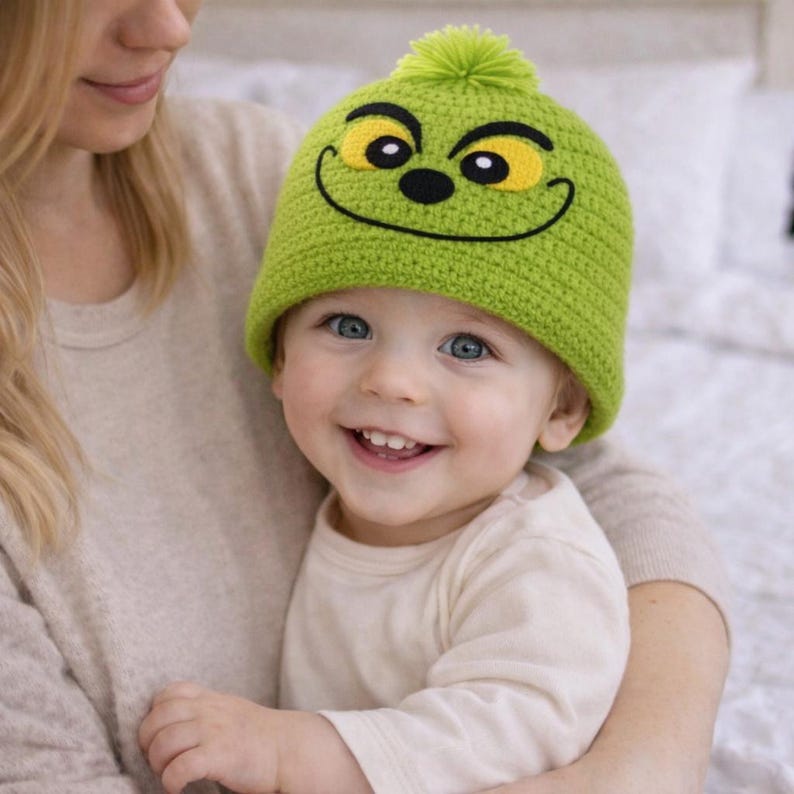Peut inclure: Un bonnet vert en tricot avec un motif de visage souriant, avec des yeux jaunes et un contour noir. Le chapeau a un pompon vert sur le dessus. Le chapeau est port&eacute; par un b&eacute;b&eacute; aux yeux bleus et une chemise blanche &agrave; manches longues.