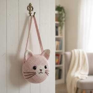 Puede incluir: Un bolso rosa con forma de gato tejido a ganchillo, con una correa larga, colgado de un gancho decorativo. El bolso presenta ojos negros, bigotes marrones y orejas con puntas blancas. El bolso est&aacute; hecho de hilo suave y texturizado.