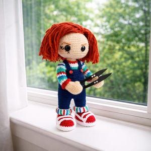 May include: Crocheted doll with red hair, blue overalls, and striped shirt. The doll is holding black scissors and wearing red and white sneakers. The doll is standing on a white windowsill with a green background.