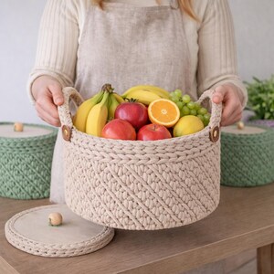 May include: A beige crochet basket filled with fresh fruit, including bananas, apples, an orange, grapes, and a lemon. The basket has two handles and is held by a person. A matching basket with a lid is in the background.