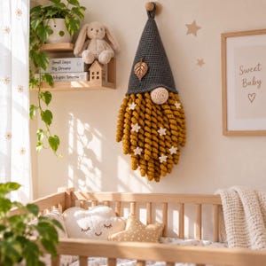 May include: A wall-hanging crochet gnome decoration featuring a dark grey pointed hat adorned with a metallic leaf charm, a round beige nose, and a long, thick beard made of mustard yellow spiral yarn curls decorated with small white crochet stars.