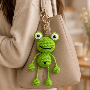 May include: A bright green crocheted frog keychain featuring a round head with large white and black eyes, a stitched smile, and a round body with a black X-stitch detail. The frog is attached to a gold-toned metal lobster clasp and ring.