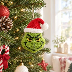 Puede incluir: Un adorno de ganchillo con forma de cabeza de personaje verde con expresión de enfado, ojos amarillos y detalles bordados en negro. El personaje lleva un gorro de Papá Noel de ganchillo rojo y blanco con un pompón blanco, colgado de un hilo.