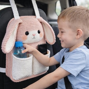May include: A pink and white crochet bunny car organizer with pockets for storage. The organizer has a bunny face with black eyes, pink cheeks, and long ears. A water bottle and a phone are in the pockets.