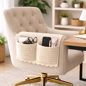Puede incluir: Organizador de brazo de silla de ganchillo color crema con dos bolsillos. El organizador está colocado sobre un reposabrazos de silla tapizado beige. Los bolsillos contienen un cuaderno, gafas, un teléfono inteligente, un bolígrafo y un tubo de loción. La silla tiene una base dorada.