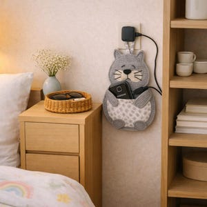 Könnte beinhalten: Ein grauer, katzenförmiger Wand-Handyhalter mit einem weiß-grau gemusterten Bauch, der ein schwarzes Handy mit einem Ladekabel hält. Der Halter ist an einer Steckdose angeschlossen. Ein geflochtener Korb mit Sonnenbrille und eine kleine Vase mit Blumen stehen auf einem Nachttisch aus Holz.