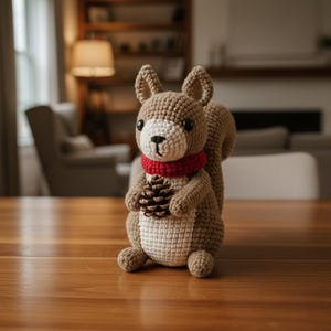 May include: A handmade crochet squirrel with a brown body, cream-colored belly, and a red scarf. The squirrel is holding a pinecone and has black eyes. The plush toy is sitting on a wooden surface.