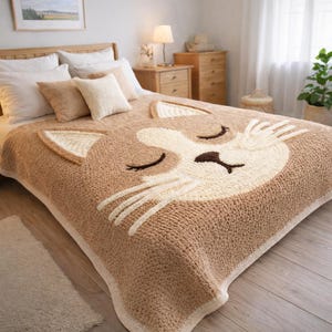 May include: A tan and cream-colored knitted throw blanket with a cat face design. The blanket features a large cat face with closed eyes, whiskers, and ears. The blanket is on a bed with pillows.