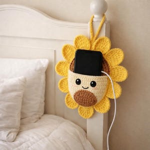Puede incluir: Un soporte para teléfono de girasol tejido a mano en colores amarillo, marrón y crema. El soporte para teléfono tiene una cara sonriente y un bolsillo para guardar un teléfono. Un cable de carga blanco está enchufado al teléfono.