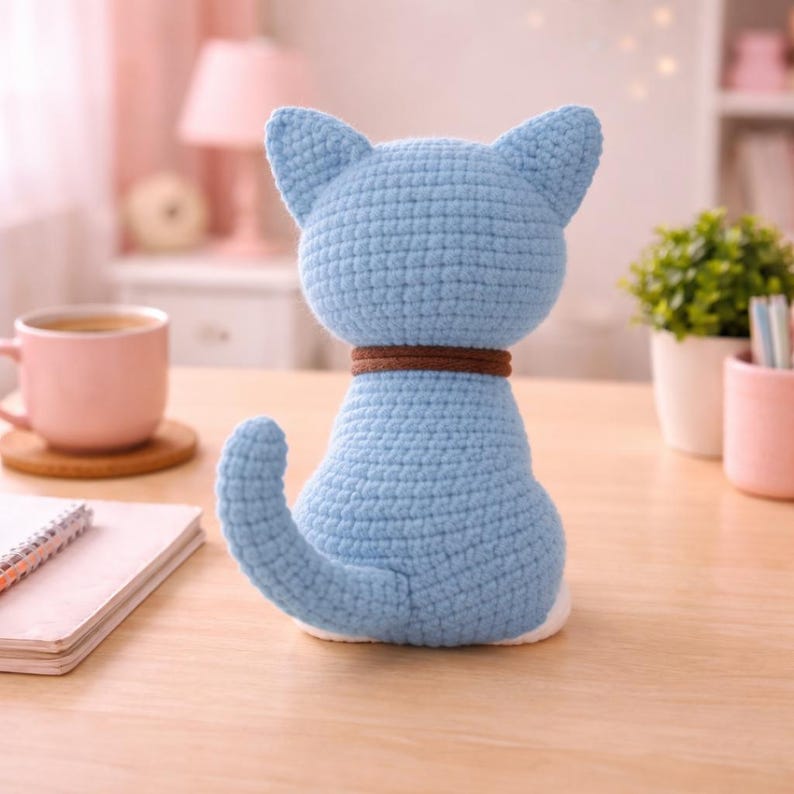 May include: A light blue crocheted cat figurine with a brown collar. The handmade cat has pointed ears and a curled tail. The cat is sitting on a wooden surface. The cat is a decorative item.