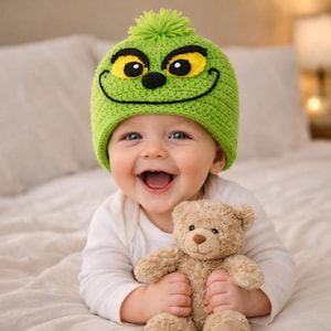 May include: A smiling baby wearing a green knitted hat with a cartoon face and a green pom-pom. The baby is holding a small brown teddy bear and wearing a white long-sleeved shirt. The background is a soft, neutral color.