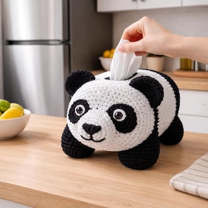 Peut inclure: Un cache-boîte à mouchoirs en forme de panda, fait au crochet, noir et blanc. Le panda a des oreilles, des yeux et des pieds noirs, avec un corps blanc. Une main tire un mouchoir de l'ouverture supérieure. Le cache-boîte est sur une surface en bois.