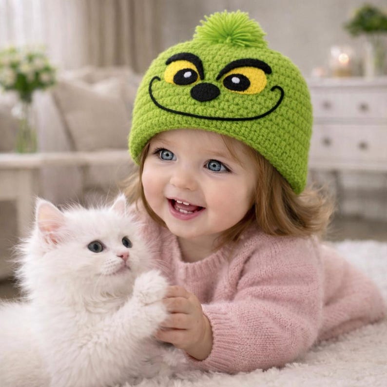 Peut inclure: Un enfant souriant porte un bonnet vert en tricot avec un visage de dessin anim&eacute; et un pompon vert. L'enfant porte un pull rose et tient un chaton blanc et duveteux. L'enfant a les yeux bleus et est allong&eacute; sur un tapis blanc.