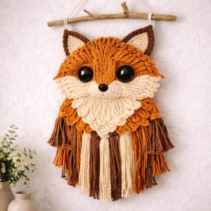 May include: A handcrafted wall hanging featuring a fox design. The fox is made of orange, brown, and cream-colored yarn, with large black eyes and a small black nose. The fox is suspended from a wooden branch.