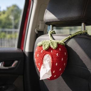 May include: A red crochet strawberry-shaped tissue holder with white embroidered seeds and a green leafy top. The holder features a central opening for dispensing white tissues and includes two green crochet straps for hanging on a car headrest.