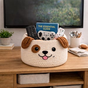 May include: A cream-colored, crocheted dog-shaped basket with brown ears and accents. The basket holds a book titled "The Essential Guide", a game controller, and two remote controls. The basket is sitting on a wooden surface.