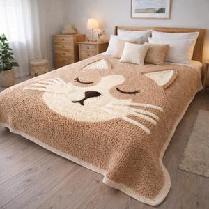 May include: A tan and cream-colored crochet blanket featuring a large cat face design. The blanket has a textured appearance with a cream-colored border and cat face details, including closed eyes and whiskers. The blanket is displayed on a bed with pillows.