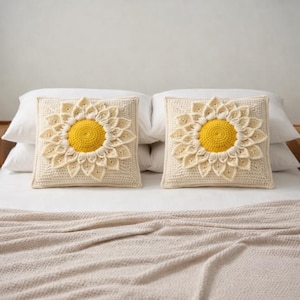 Puede incluir: Dos cojines cuadrados de crochet color crema con un gran dise&ntilde;o de girasol. El centro del girasol es amarillo, rodeado de p&eacute;talos color crema. Los cojines est&aacute;n sobre una cama blanca con una manta beige.