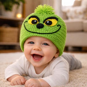 Peut inclure: Un b&eacute;b&eacute; souriant portant un bonnet vert crochet&eacute; avec un motif de visage de dessin anim&eacute;. Le chapeau pr&eacute;sente des yeux jaunes, un nez noir et une bouche souriante. Le b&eacute;b&eacute; porte un t-shirt blanc &agrave; manches longues et un pantalon gris, couch&eacute; sur un tapis clair.