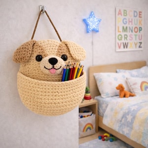 May include: A beige crocheted dog-shaped wall organizer with a basket for storage. The dog has brown ears and a black nose and eyes. The basket is filled with colored pencils. A brown rope is used for hanging.