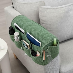 May include: A green fabric sofa arm organizer with multiple pockets. The organizer is filled with items like a water bottle, notebook, pens, glasses, and a spray bottle. The organizer is placed on the arm of a gray sofa.