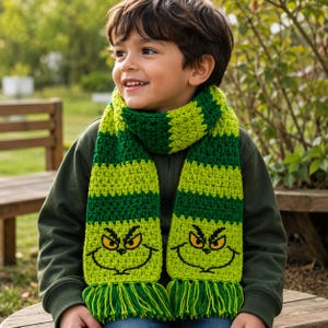 May include: A crochet scarf featuring alternating horizontal stripes of lime green and dark forest green yarn. The scarf ends display a stitched graphic of a yellow-eyed, frowning character face with black outlines and a fringe of green yarn.