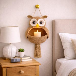 May include: A handcrafted, brown owl-shaped wall hanging with a candle holder. The owl features large white and black eyes, small ears, and a tassel. A lit candle sits inside the woven basket. The owl is suspended from a gold-colored hook.
