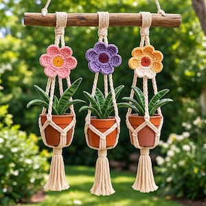 Puede incluir: Un colgante de pared con tres macetas de terracota sujetas por cuerdas de macramé color crema suspendidas de una rama de madera. Cada cuerda incluye una flor de ganchillo en rosa, morado o amarillo y termina en una borla decorativa.