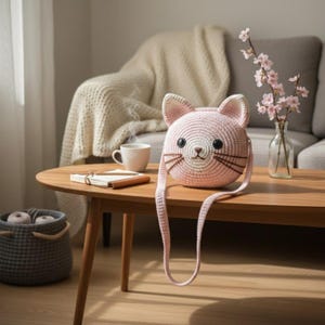 Puede incluir: Un bolso rosa y blanco con forma de gato tejido a ganchillo, con una correa larga. El bolso tiene ojos negros, bigotes marrones y orejas rosas. Est&aacute; sobre una mesa de madera con un cuaderno, una taza de caf&eacute; y un jarr&oacute;n con flores.