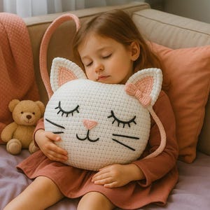Puede incluir: Una almohada blanca de ganchillo con forma de gato con detalles en rosa, incluyendo orejas y un lazo, es sostenida por una persona. La almohada presenta ojos, bigotes y nariz bordados en negro. Se ve un pequeño oso de peluche.