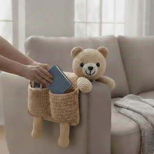 May include: A crocheted teddy bear-shaped organizer in beige, designed to hang over the arm of a sofa. The bear has a smiling face and two pockets for storage. A person is placing a blue book into one of the pockets.