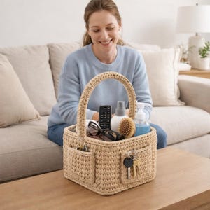 May include: A beige crochet basket with a handle, two side pockets, and a rectangular shape. The basket contains a remote control, glasses, a brush, and bottles. Keys hang from one pocket. The basket is on a wooden table.