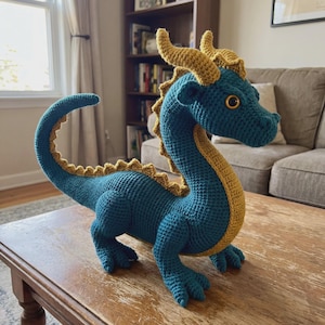May include: A teal and gold crochet dragon figurine. The dragon has a long tail, horns, and a spiky spine. The dragon is sitting on a wooden table. The dragon's belly and the inside of its neck are gold.