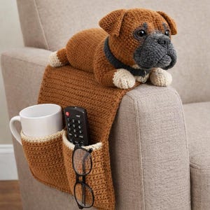 Pode incluir: Um organizador de poltrona de crochê marrom com bolsos para uma caneca, controle remoto e óculos. Um cão boxer de crochê repousa em cima. O organizador é projetado para caber no braço de uma cadeira.