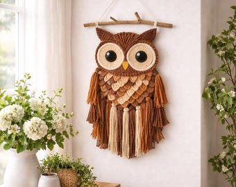 May include: A handcrafted owl wall hanging made of brown, tan, and cream-colored yarn. The owl has large black eyes, a yellow beak, and decorative tassels. It is suspended from a wooden branch with a string.