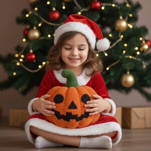 Op de afbeelding: Een kind met een rode kerstmuts en jurk houdt een oranje gehaakte pompoen vast met een groene steel. De pompoen heeft zwarte ogen, een neus en een mond. Op de achtergrond een kerstboom met ornamenten.