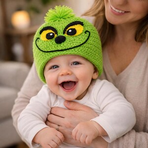 Peut inclure: Un chapeau de b&eacute;b&eacute; vert crochet&eacute; avec un motif de visage du Grinch, avec des yeux jaunes, un nez noir et une bouche souriante. Le chapeau a un pompon vert sur le dessus. Le b&eacute;b&eacute; porte une chemise blanche &agrave; manches longues.