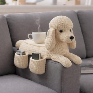 Può includere: Un bracciolo per divano a forma di barboncino all'uncinetto, color crema, con due tasche e una superficie piana per una tazza. Il barboncino ha occhi neri e naso nero. Una tazza bianca con vapore è appoggiata sulla superficie piana.