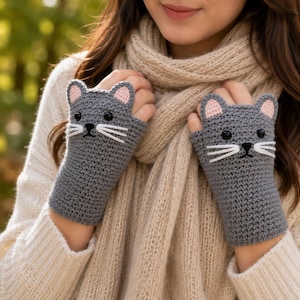 May include: A pair of gray fingerless gloves crocheted in a cat design. Each glove features two small ears with pink centers, black bead eyes, and three white embroidered whiskers on each side of a small black stitched nose and mouth.