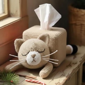 Op de afbeelding: Een beige gehaakte tissueboxhoes in de vorm van een slapende kat. De kat heeft gesloten ogen, witte snorharen en een witte en bruine neus. Witte tissues komen uit de bovenkant. De staart van de kat is zwart en wit.
