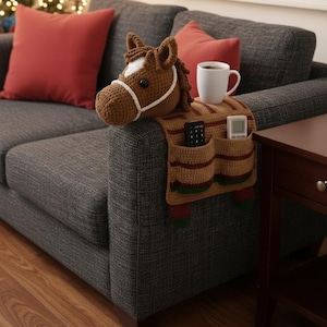 Puede incluir: Una funda de reposabrazos con cabeza de caballo de ganchillo marrón con una mancha y brida blancas, y un diseño de bolsillo a rayas. La funda tiene bolsillos para mandos a distancia y una taza de café.