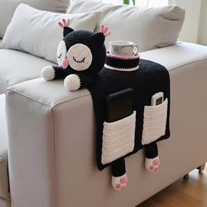 Puede incluir: Organizador de reposabrazos de sofá con forma de búho, tejido a ganchillo en blanco y negro. Cuenta con un portavasos, dos bolsillos y detalles de búho como ojos, orejas y patas. Una taza blanca con borde rosa está en el portavasos.