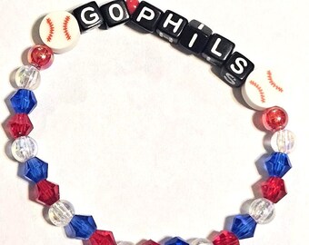 May include: A beaded bracelet with the word "GOPHILS" spelled out in black letter beads. The bracelet features red, white, and blue beads, along with baseball-shaped beads. The bracelet is a patriotic accessory.