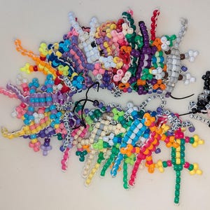 May include: A collection of colorful beaded lizard and abstract figures. The figures are made of small, brightly colored beads in various shapes and sizes. Some figures have names spelled out in white beads. The figures are arranged on a white surface.