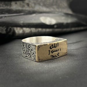 May include: A silver and gold ring with intricate filigree detailing on the sides. The gold rectangular face of the ring features Arabic script. The ring is set against a dark, textured background.