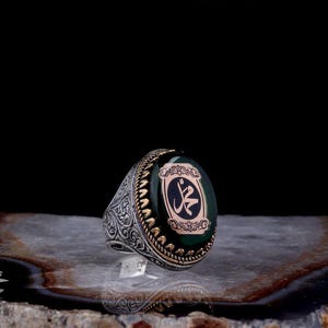 May include: A silver ring with an oval, dark green stone. The stone features a gold-colored emblem with Arabic script. The ring has intricate, engraved detailing on the band and a gold-colored border around the stone.