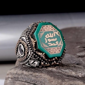 May include: A detailed silver ring with an octagonal green stone. The stone has gold lettering and ornate gold detailing. The ring has a textured band with black accents and a black stone on the side. The ring is on a gray stone surface.