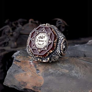 May include: A silver ring with a dark red center stone. The center stone has a gold-colored star design with Arabic script. The ring has a detailed, textured band with additional gold accents. The ring is displayed on a dark, textured surface.