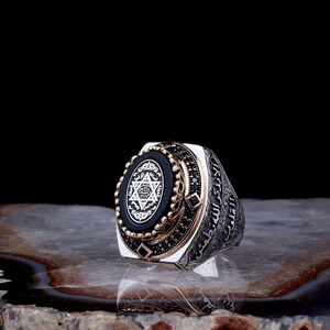 May include: A silver ring with an oval black onyx center stone featuring a white Star of David design. The ring has gold accents, black gemstones, and Arabic script detailing the band. The ring is displayed on a stone surface.